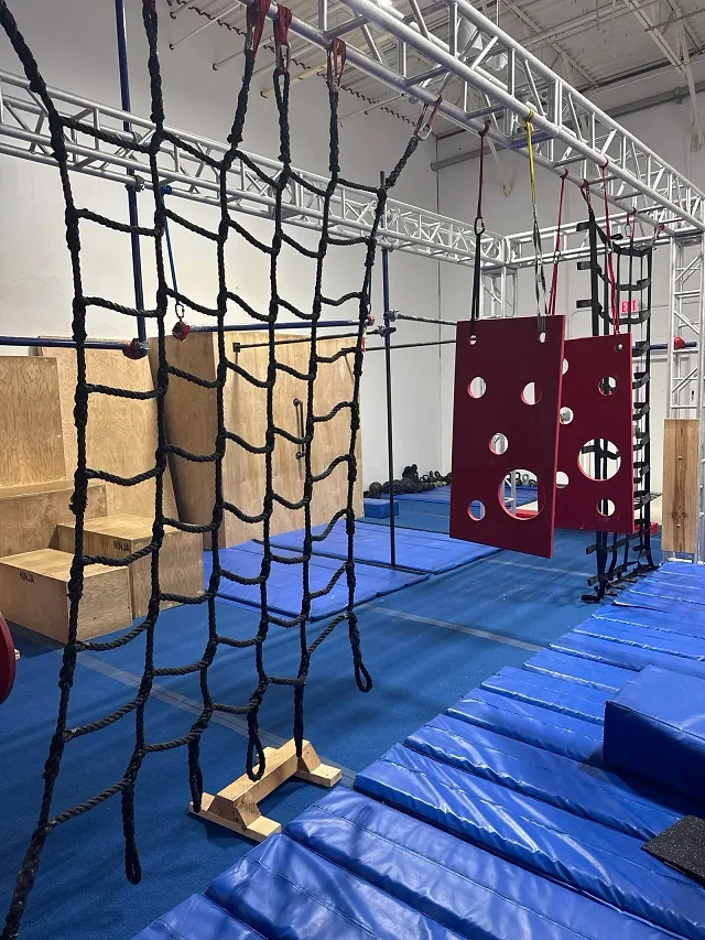 A rope cargo net hanging from the steel rig, with red peg boards, a strap ladder, plyo boxes and blue mats behind it
