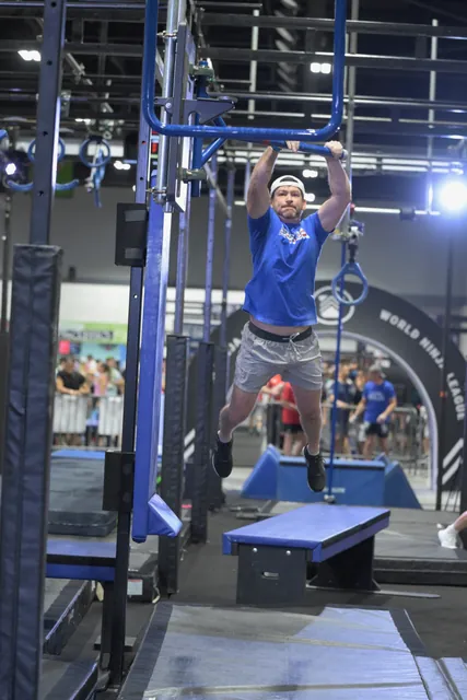 David Kiser in a blue Team Ninja shirt swinging from a bar in front of the World Ninja League arch