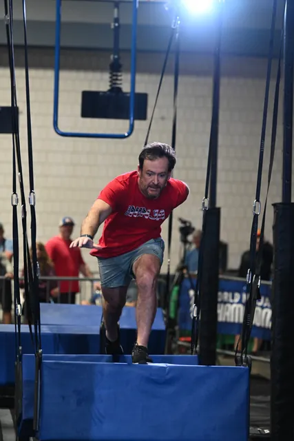 David Kiser in a red Team Ninja shirt lunging across hanging steps