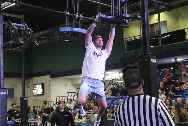 David Kiser hanging from a spinning obstacle as a referee and the crowd watch