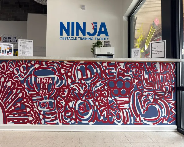 The NinjaFit front desk under the NINJA Obstacle Training Facility sign, its counter covered in a hand-painted red, white and blue mural with trophies and the words We Do Ninja