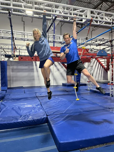Nicholas Parker and Jessie Graff hanging one-handed from a bar on the NinjaFit course, both smiling
