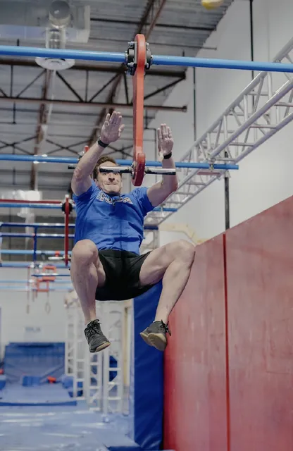 Nicholas Parker in a blue Team Ninja shirt leaping for a swinging handle on the NinjaFit course