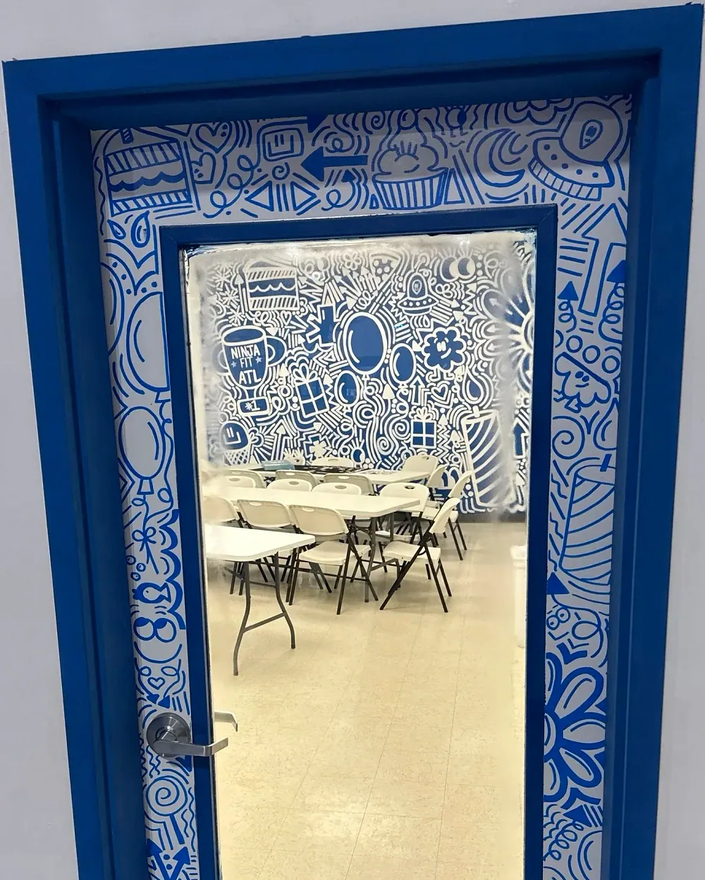 The blue party room's door, hand-painted with blue doodles, with the mural, tables and chairs visible through its window