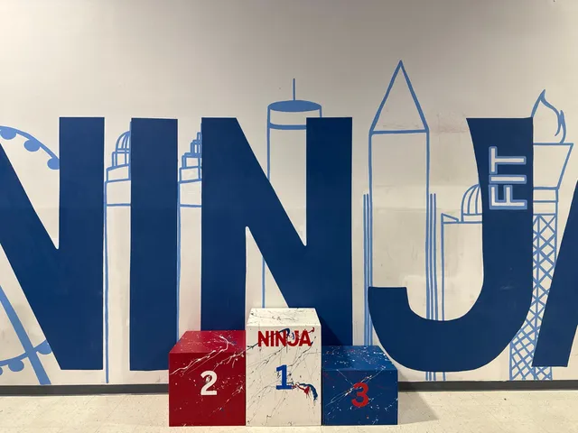 A paint-splattered winners' podium: a red 2, a white 1 lettered NINJA and a blue 3, against the blue NINJA FIT skyline mural