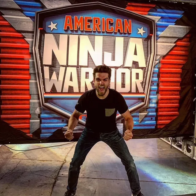 Tyler Gillett flexing and shouting in front of the American Ninja Warrior sign