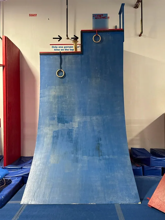 NinjaFit's tall blue stepped warped wall with rings at the top, a sign reading Only one person at a time on the top, and the 14-Foot Achievers board above it