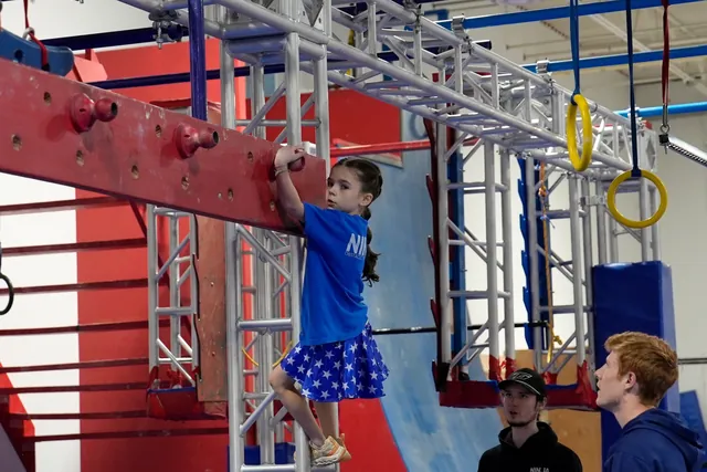 Elliot in a star skirt climbing a ladder to the peg board while two people watch from below